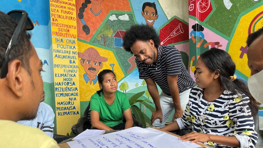 Six people collaborate around a piece of paper on a table in front of a wall with brightly coloured illustrations of people.