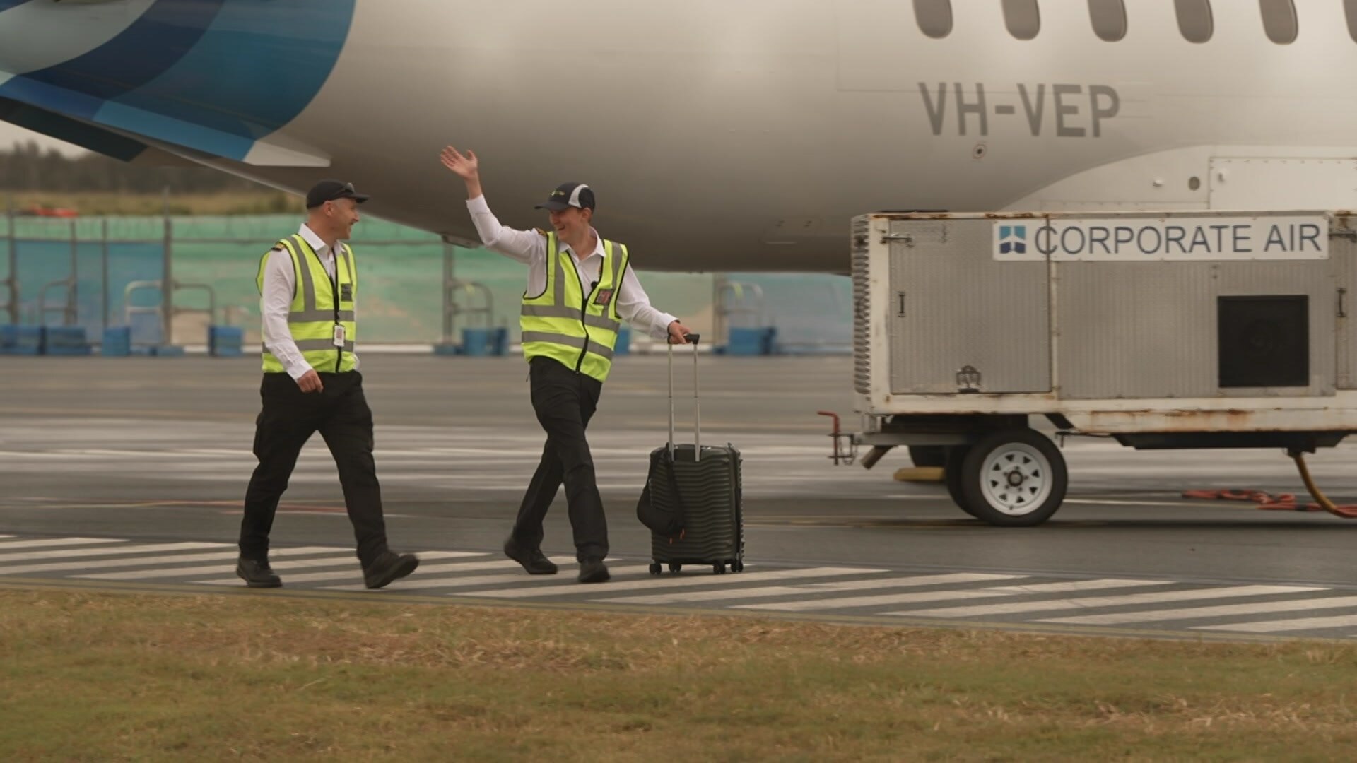 Two pilots on the tarmac in high vis yellow vests, one waves