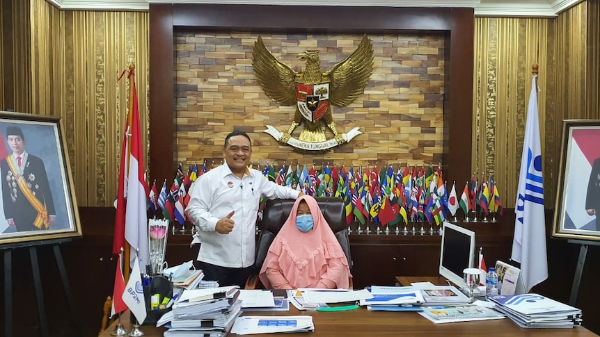 A woman sits in a chair wearing a mask and a pink hijab while a man stands next to her.