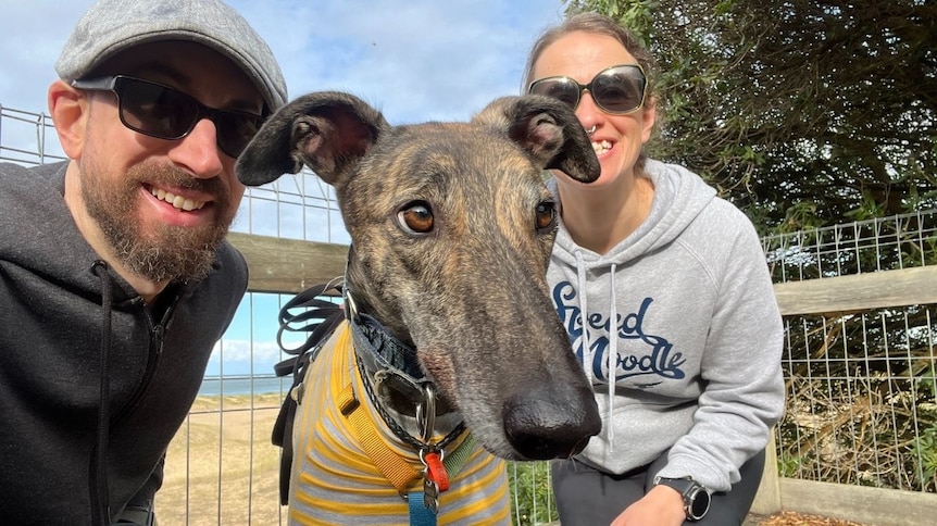 Travis Evans wears a black T-shirt and his partner Nerida wears a grey hoodie. They are posing for a selfie with a greyhound.