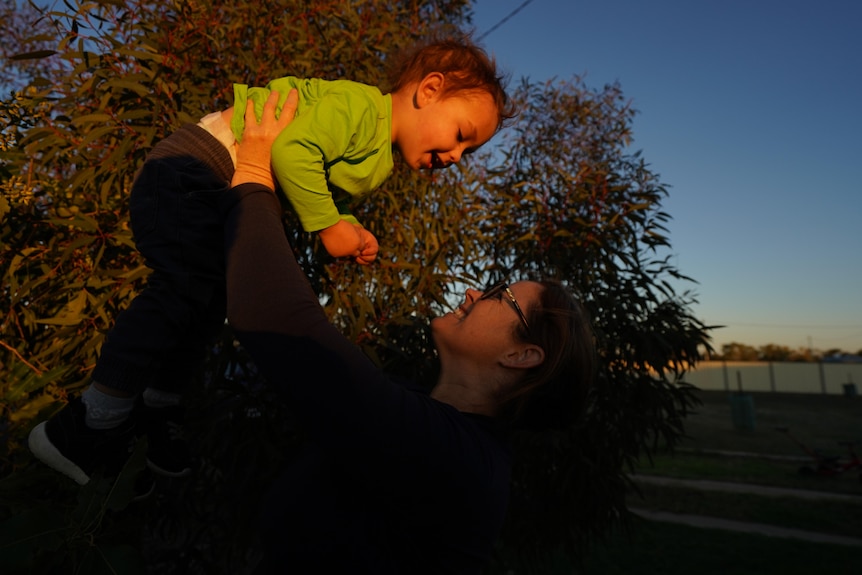 Woman holding two-year-old boy above her face, smiling, in garden with sunset in background.