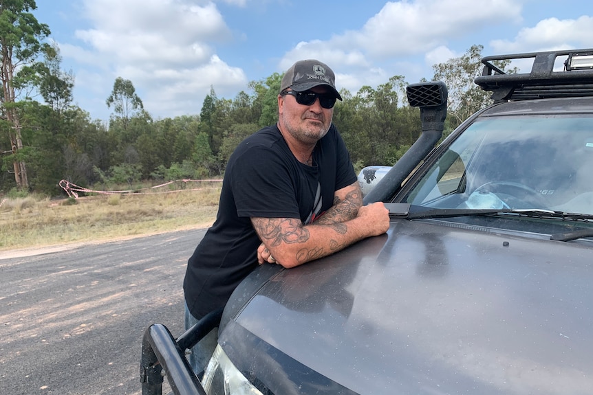 a man wearing a black t shirt, sunglasses and a cap leans on the bonnet of his four wheel drive