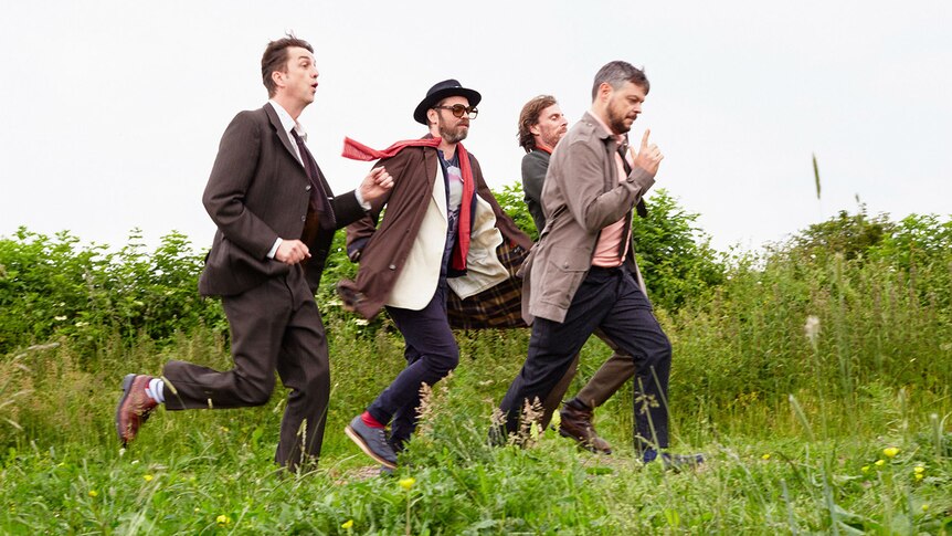 The four members of Supergrass run through a green field while wearing suits.
