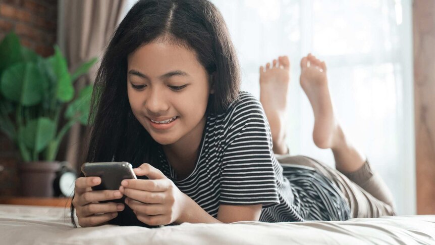 A young, diverse teenager is on her bed, lying on her front, and smiling as she looks at her phone.