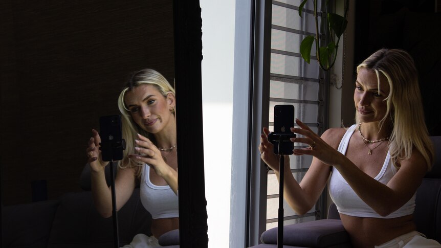 A young woman positions her phone on a stand while sitting beside a mirror