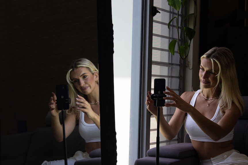 A young woman positions her phone on a stand while sitting beside a mirror