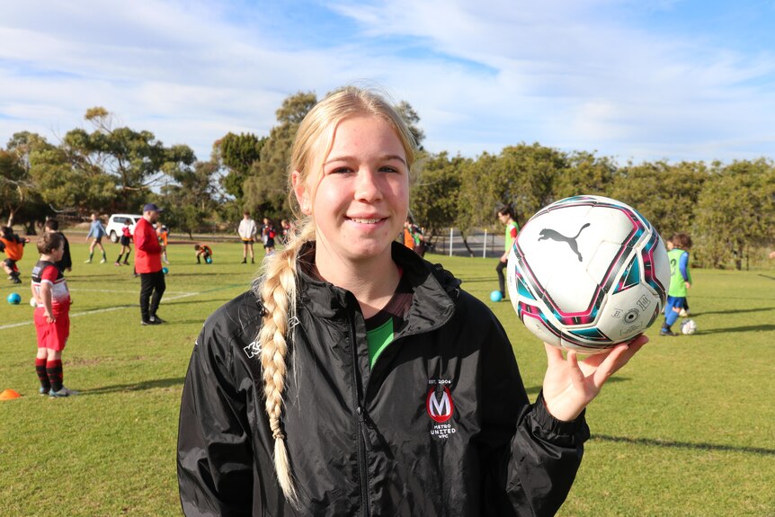 Girl with long blonde hair in plait holds a soccer ball on and oval and smiles at camera.