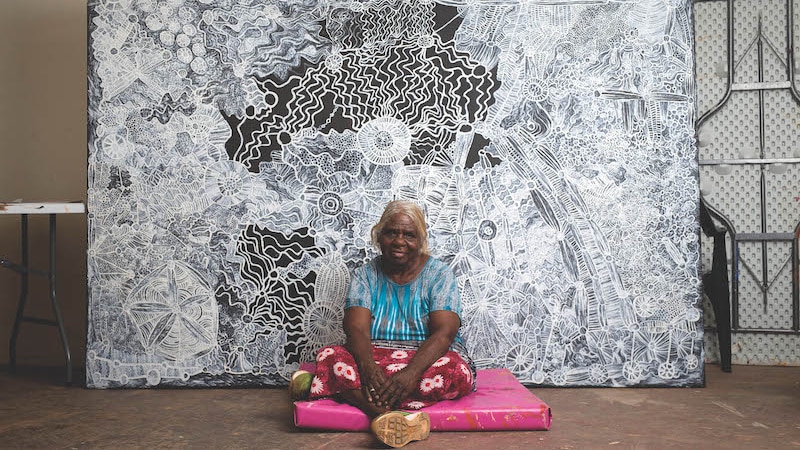 An older Aboriginal woman in colourful clothes sits in front of a vast detailed canvas painting in black and white
