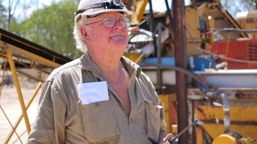 Kinglsey Fancourt, white hard hat, head lamp, glasses, holds a two-way radio, khaki shirt, yellow mining plant machinery behind.