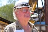 Kinglsey Fancourt, white hard hat, head lamp, glasses, holds a two-way radio, khaki shirt, yellow mining plant machinery behind.