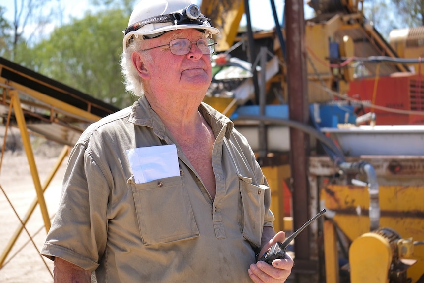 Kinglsey Fancourt, white hard hat, head lamp, glasses, holds a two-way radio, khaki shirt, yellow mining plant machinery behind.