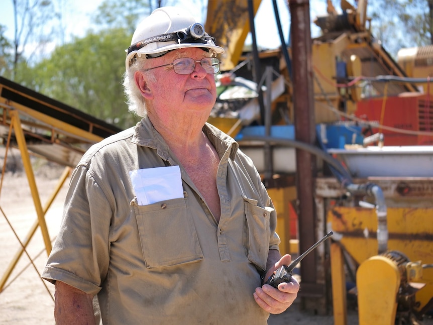 Kinglsey Fancourt, white hard hat, head lamp, glasses, holds a two-way radio, khaki shirt, yellow mining plant machinery behind.