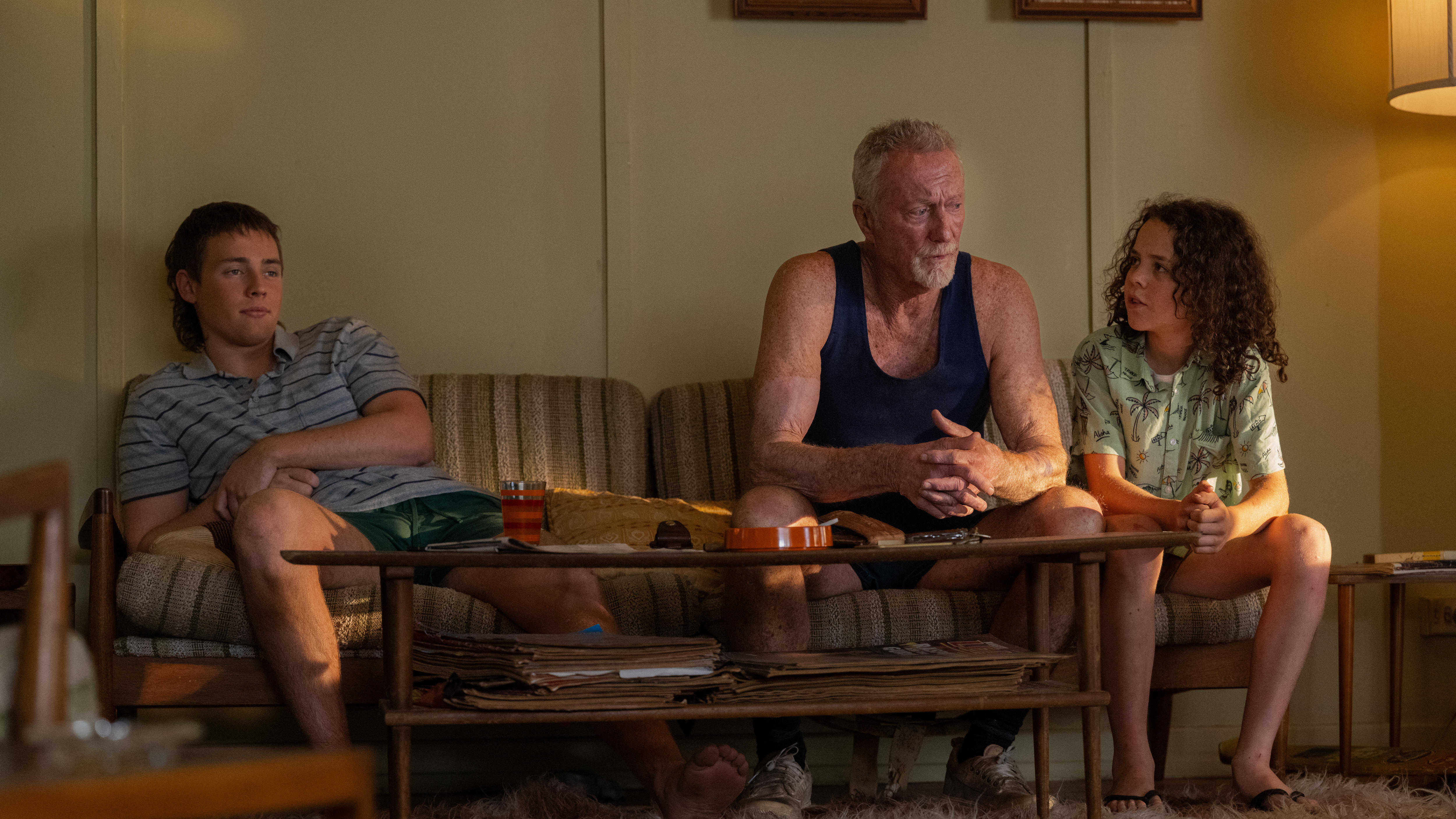 Two young boys sit on a couch with an older man in a singlet sitting in the middle watching TV.