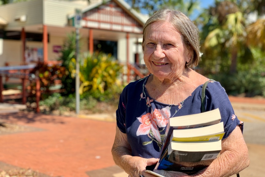 A woman holding books stands outside in the sunshine