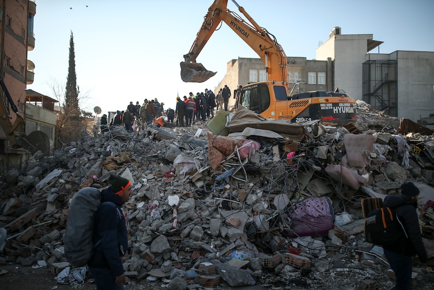 Rescue workers stand atop the rubble of a collapsed building, as earth-mover digs.