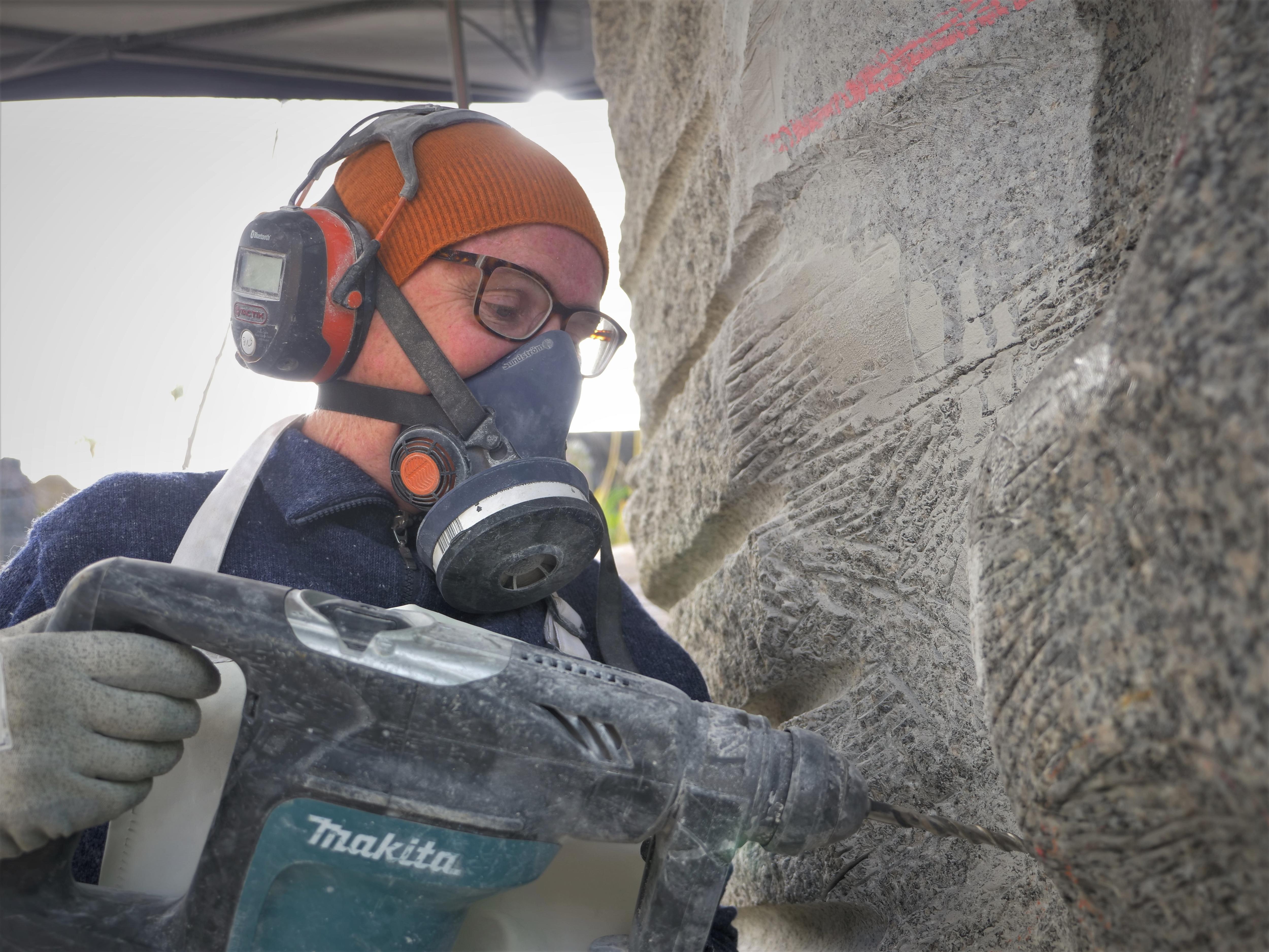 A woman wearing beanie, respirator and ear muffs drilling into a granite statue.