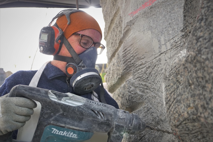 A woman wearing beanie, respirator and ear muffs drilling into a granite statue.