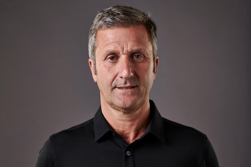 A man in a cycling team shirt stands facing a camera with his hands behind his back.