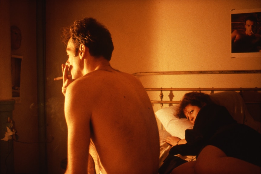 A shirtless man is perched on the edge of a bed, smoking a cigarette. A woman, fully clothed, is lying on the bed looking at him