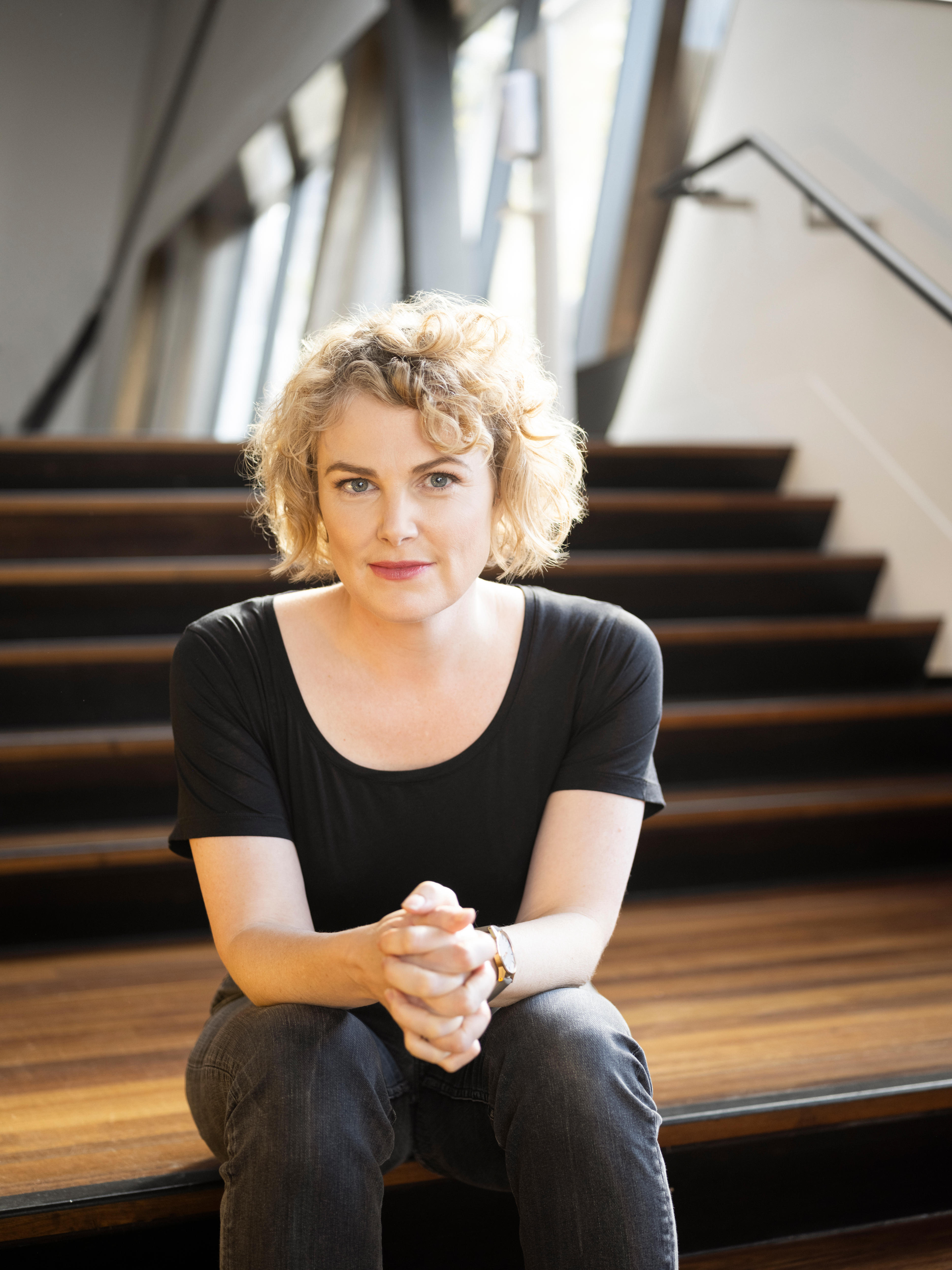 A blonde, curly-haired woman, wearing a black shirt, sits on theatre foyer stairs, hand clasped, with a slight smile on her face