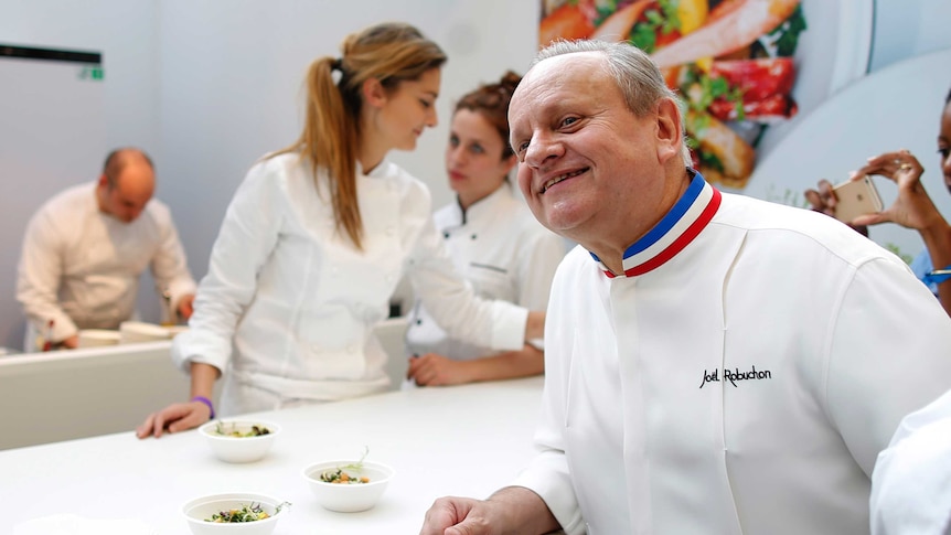 Joel Robuchon smiles for a photo with dishes of food on a table next to him.
