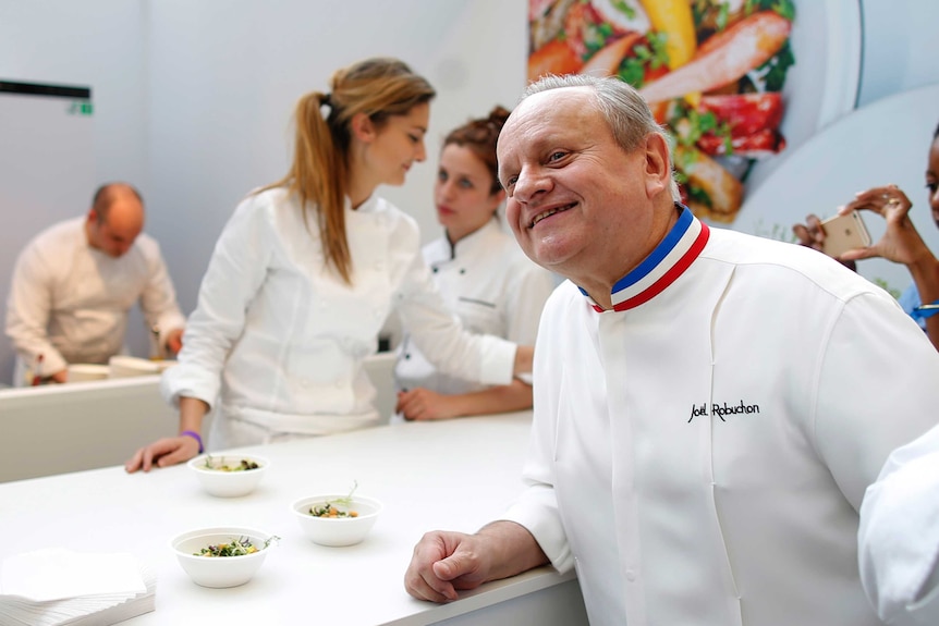 Joel Robuchon smiles for a photo with dishes of food on a table next to him.