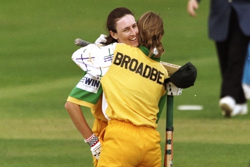 Two women cricketers embrace in the middle of the pitch