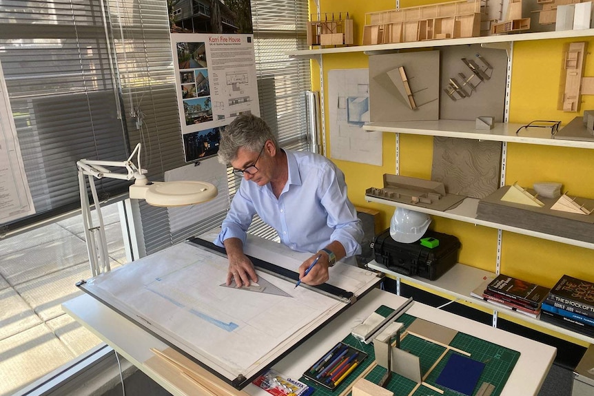 Architect Dr Ian Weir sits at a desk in an office writing and drawing on a building plan.