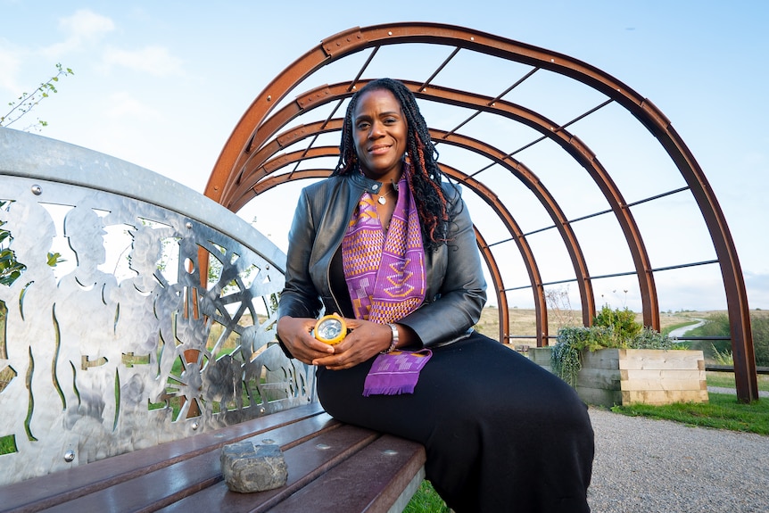 A woman with dark hair sits on a bench outside next to a piece of coal.