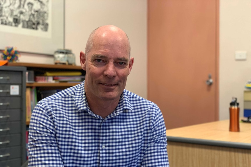 Brendan Staple, a man with a checked shirt, sits in a principal's office and looks at the camera.