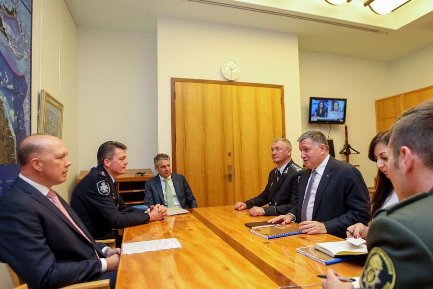 Peter Dutton, wearing suit, Andrew Colvin, wearing AFP uniform, sit at wooden table opposite men in suits in meeting room