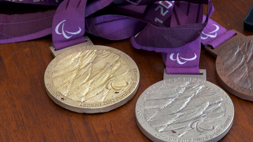 Three medals, one gold, one silver and one bronze sit on a wooden surface. They have purple ribbon and the Paralympic logo.