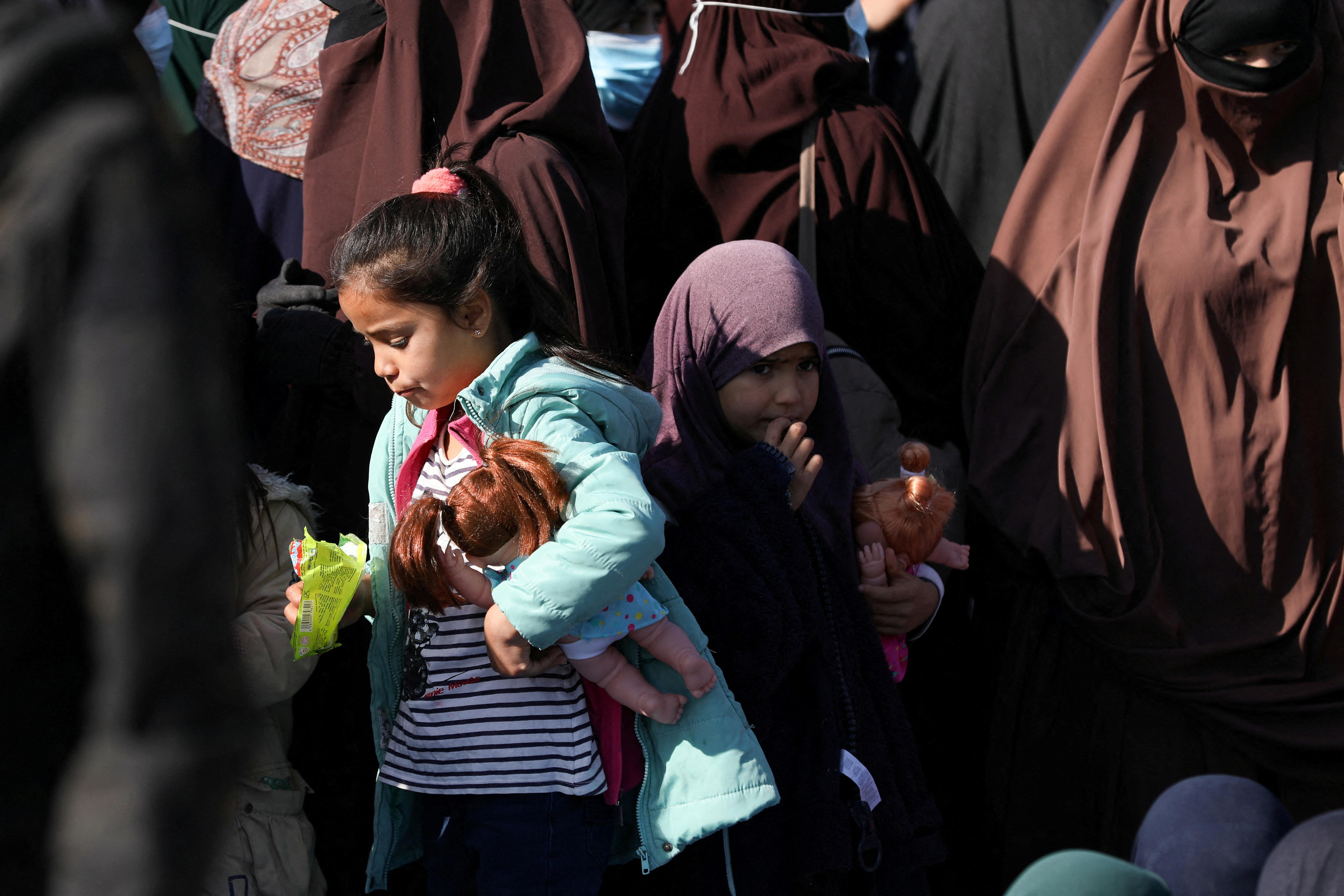 Children hold their toys while standing in front of women in brown Islamic garb.