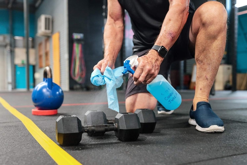 A man kneeling in a gym holds a bottle of disinfectant and sprays it on a dumbbell while he holds a rag in his other hand