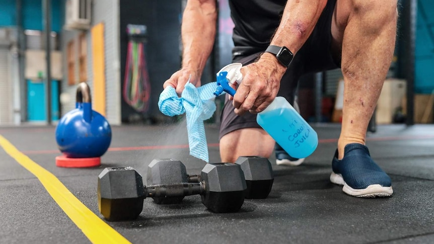 A man kneeling in a gym holds a bottle of disinfectant and sprays it on a dumbbell while he holds a rag in his other hand.