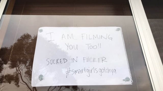 A hand-written sign stuck in a window reads: "I am filming you too, sucked in (censored)". Trees are reflected in the window.