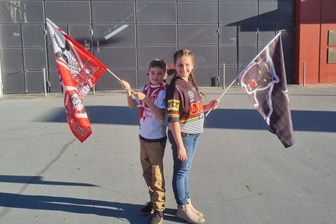 A boy and girl standing back to back, holding colourful team flags in front of a big stadium wall.
