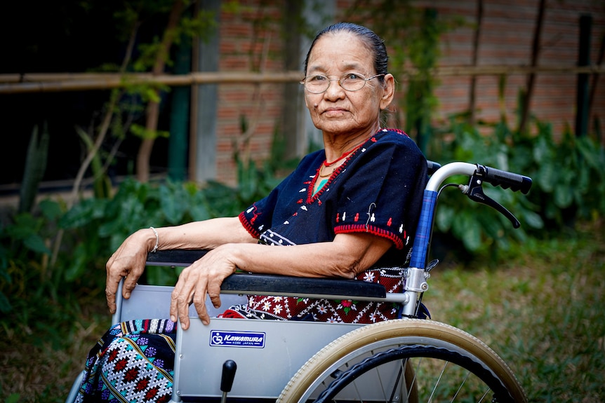 A woman sits in her wheelchair with a slight smile on her face. She is wearing traditional dress and silver-rimmed glasses.