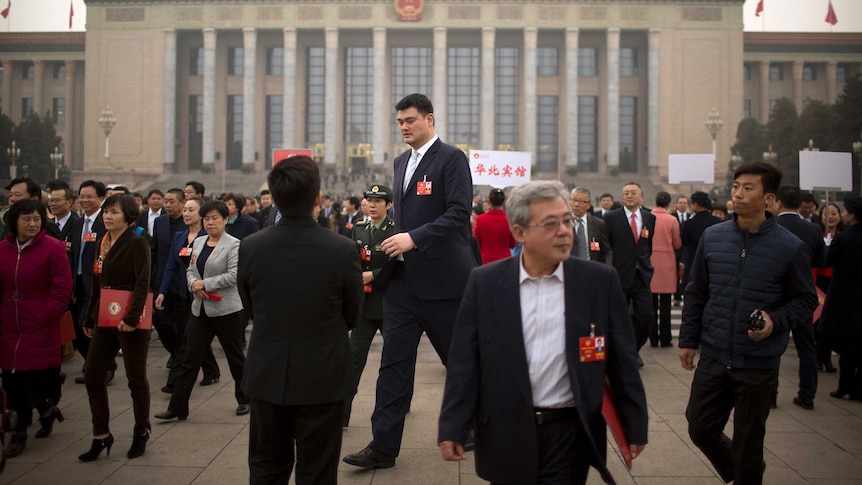 A very tall man in a suit strides through a square in Beijing, wearing an ID badge on his lapel.