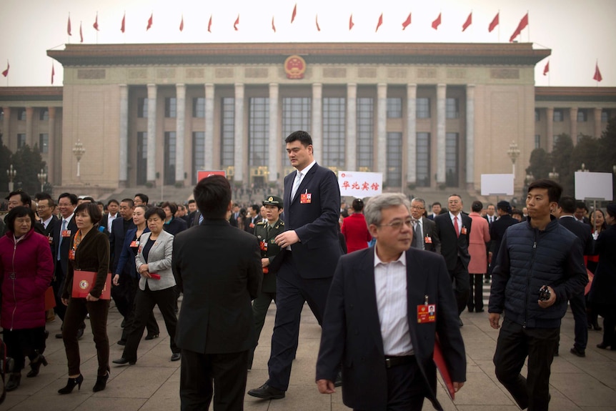 A very tall man in a suit strides through a square in Beijing, wearing an ID badge on his lapel.