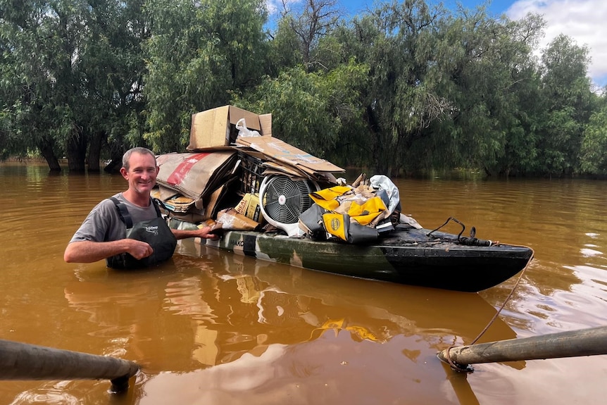 A man standing in floodwaters next to a kayak he is using to transport belongings. 