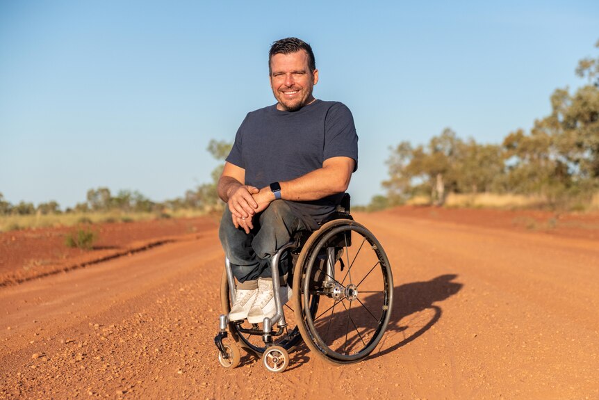 Man sitting in wheel chair in red dirt landscape on a bright blue sky day. 