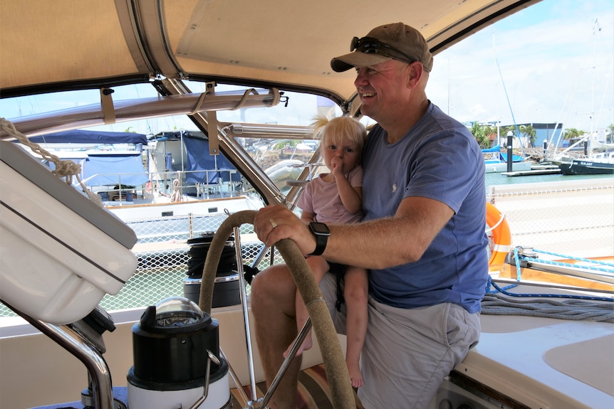 A man in his 40s at the helm of a boat, holding his one year old daughter who is sucking on her thumb. 