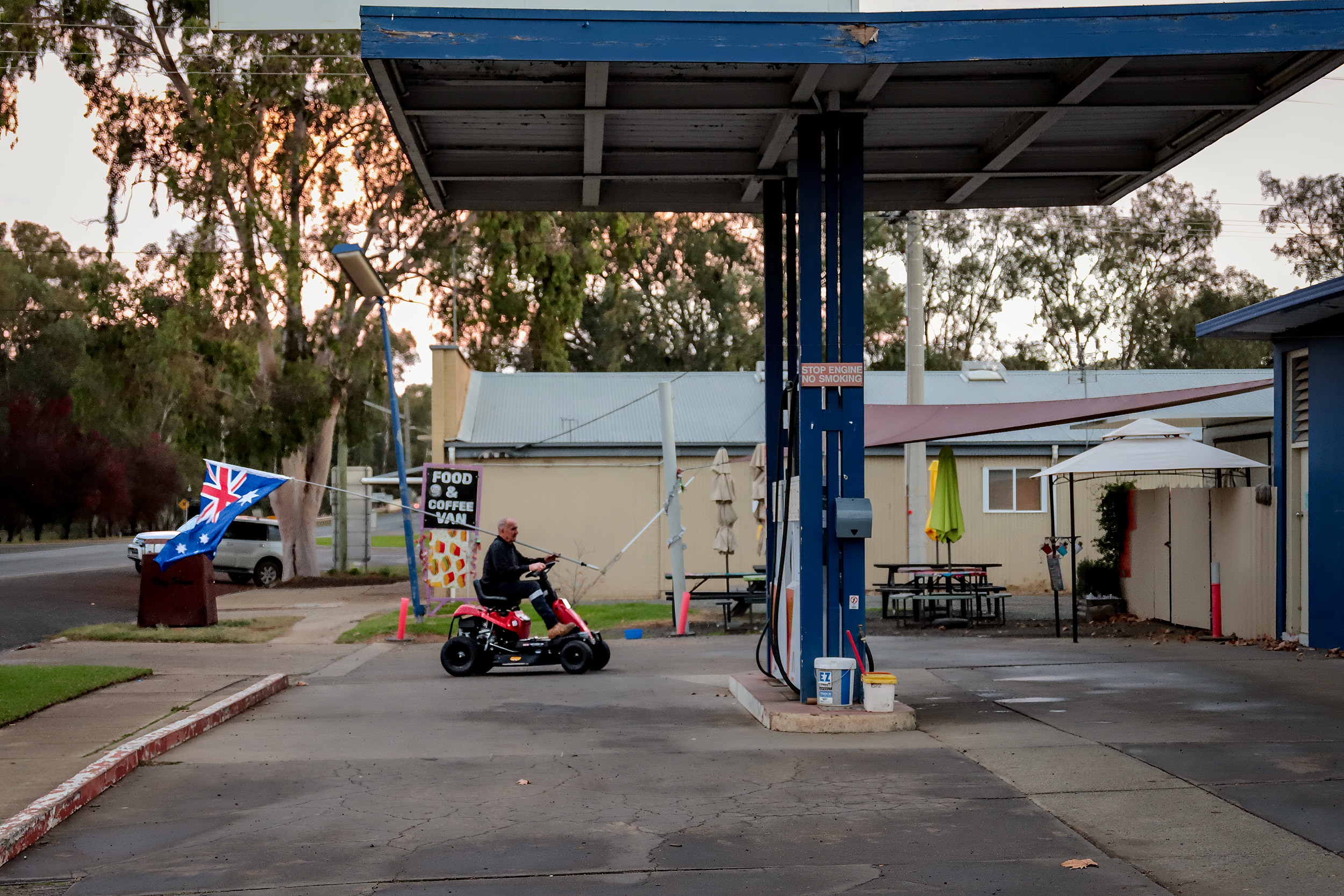 Man driving four wheel vehicle holds large Australian flag as he drives through country service station at dusk