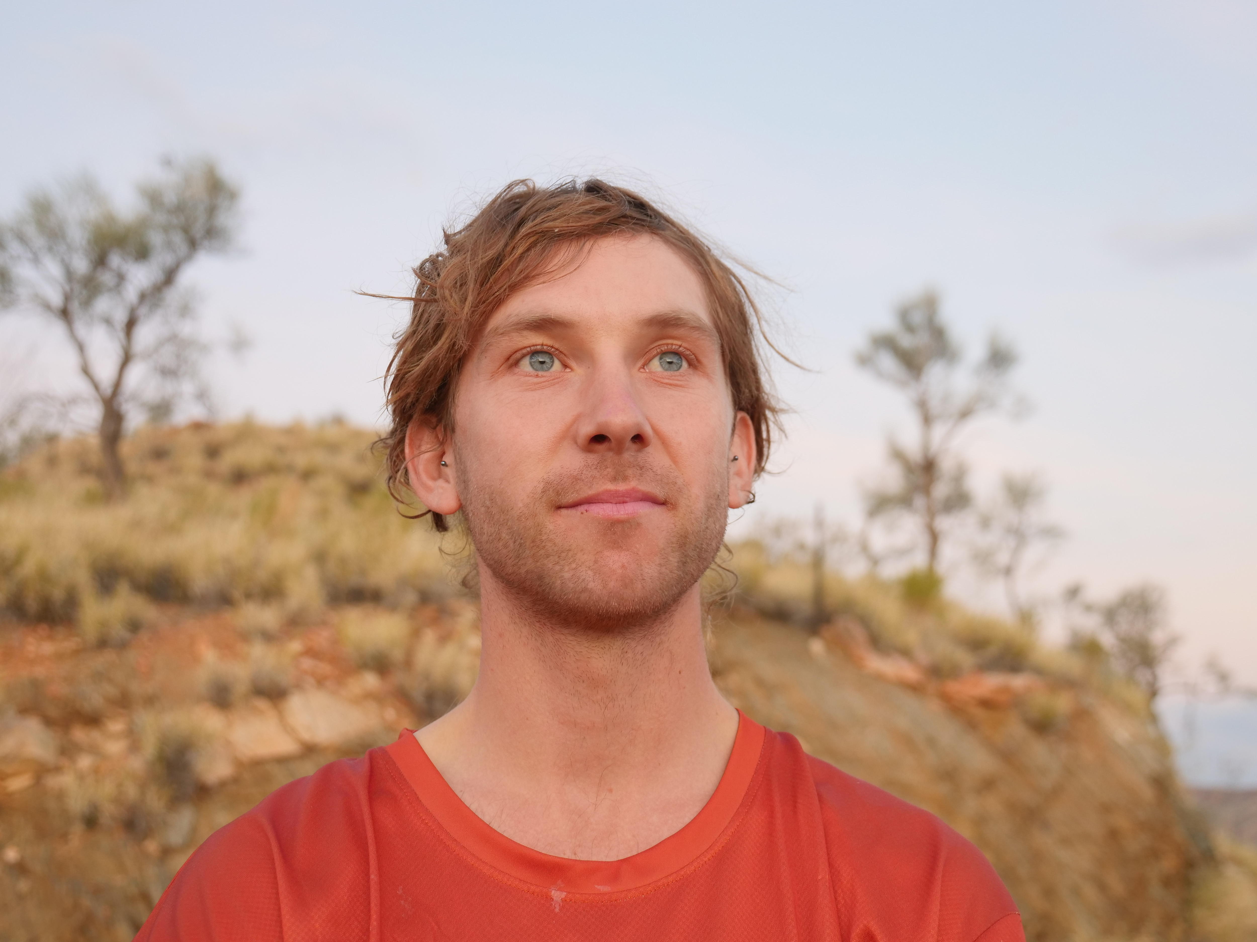 A man in an orange shirt looks up at the sky with the outback behind him.
