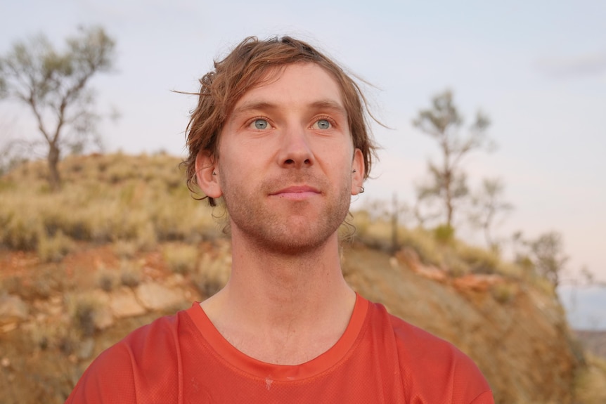A man in an orange shirt looks up at the sky with the outback behind him.