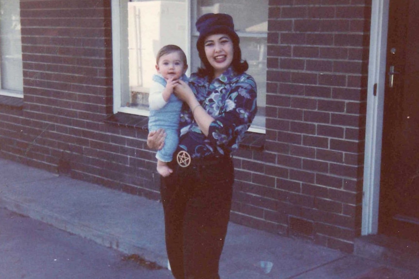 Tracey Connell smiles as she stands outside a brick house holding up her infant son.