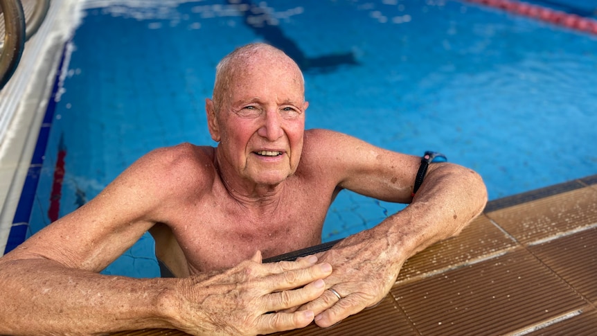 An elderly gentleman in a large public pool, leaning against the side, looking at the camera