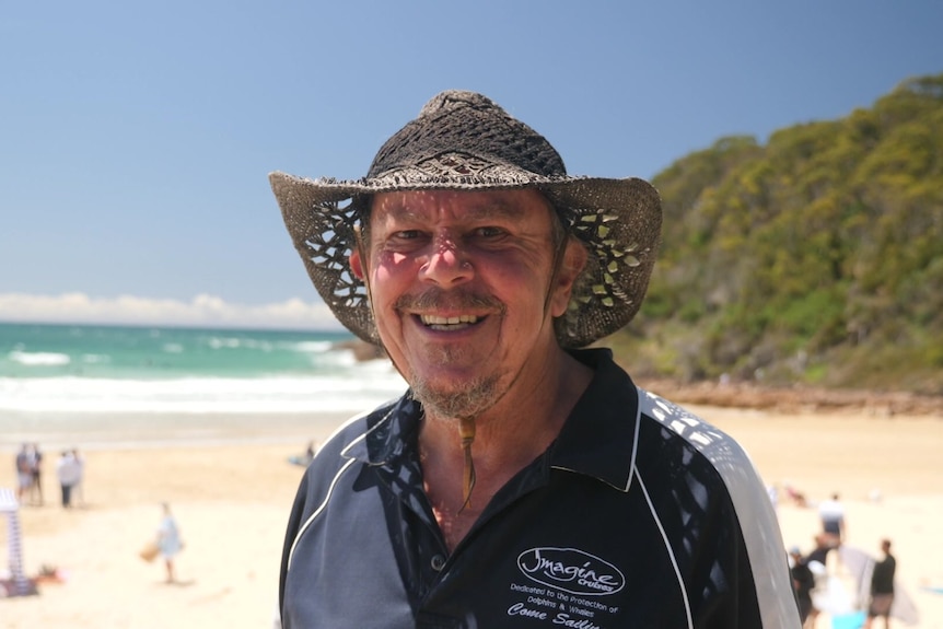 a man wearing a wicker beach hat smiling at the camera. He is standing on the sand at One Mile Beach