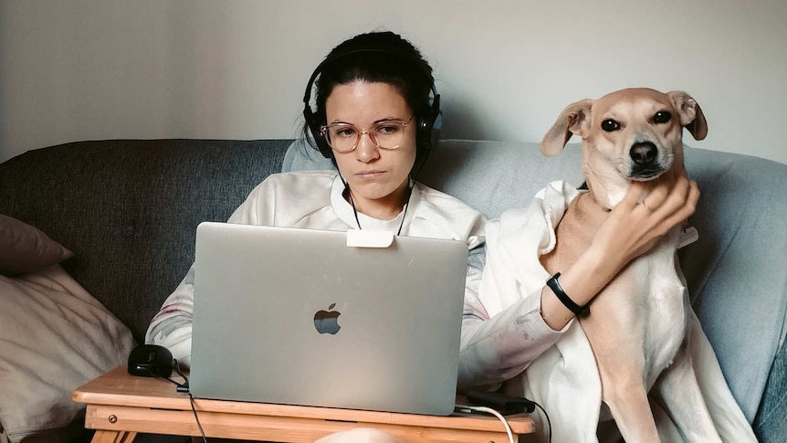 A woman with headphones pats her dog while working on a laptop